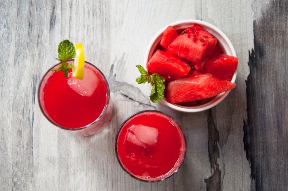 Top view of two glasses of red drink and a cup full of fruit chunks.