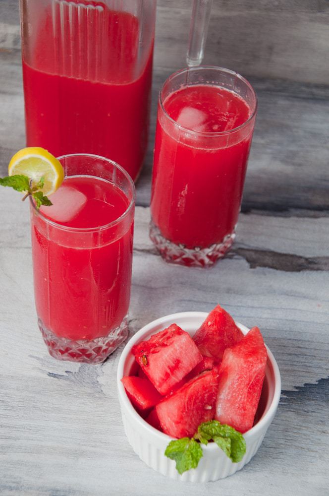 Red drink served in a pitcher with two glasses and a bowl of fresh watermelon chunks.