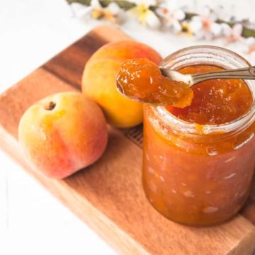 a spoon of peach jam kept of the jam jar with 2 peaches in the background.
