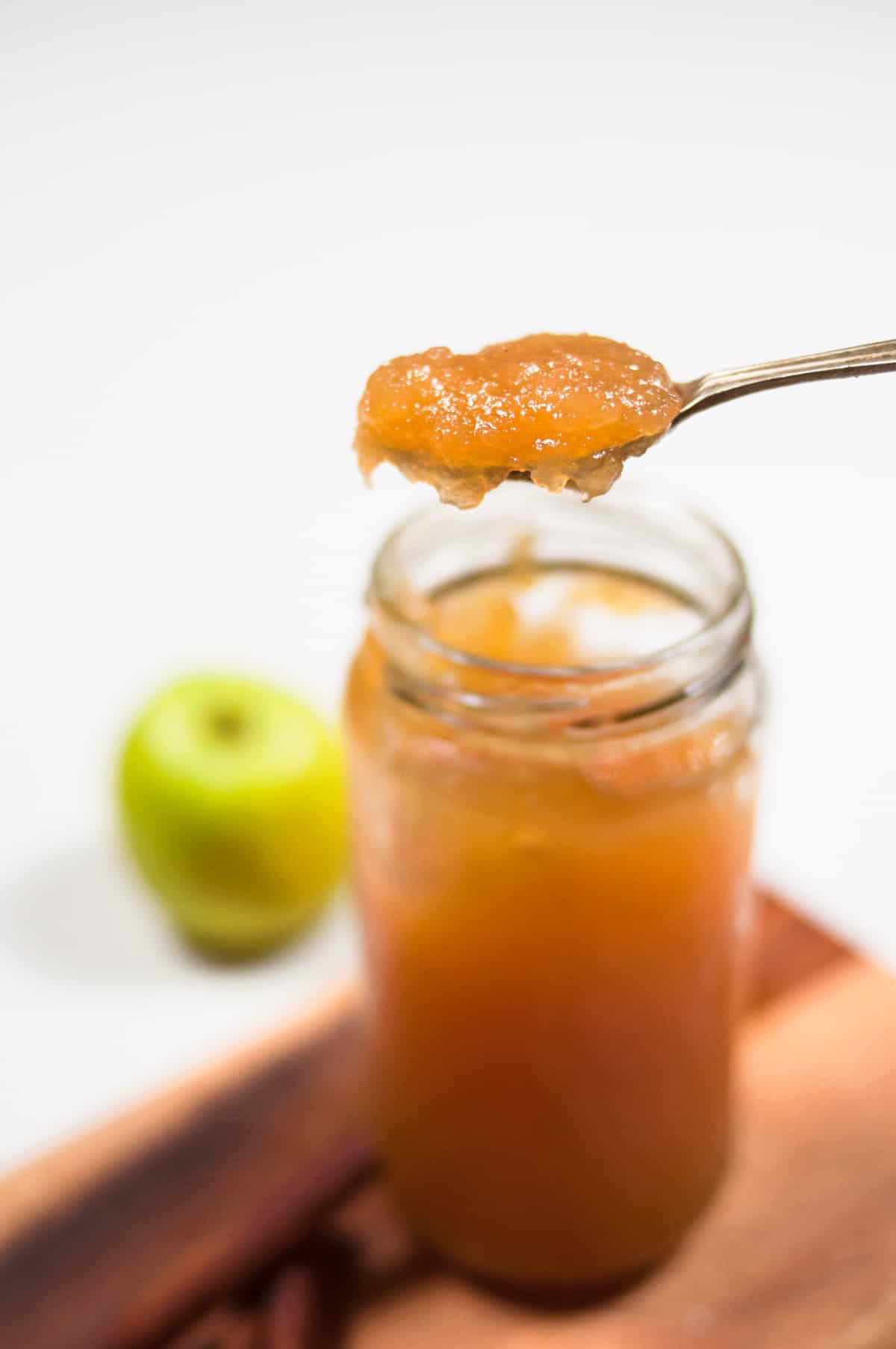 A spoon full of jam over the jar with green apple in the background.
