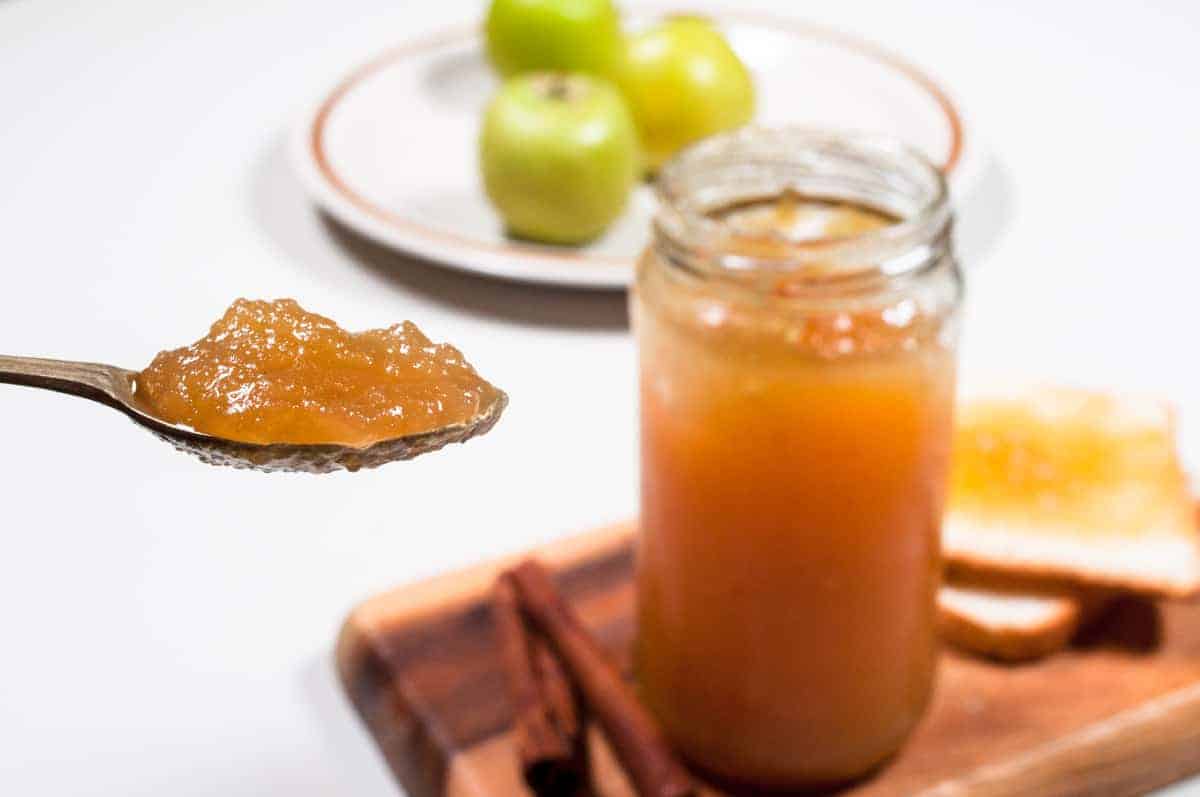 A spoon full jam in front with jam jar and apples in the background.