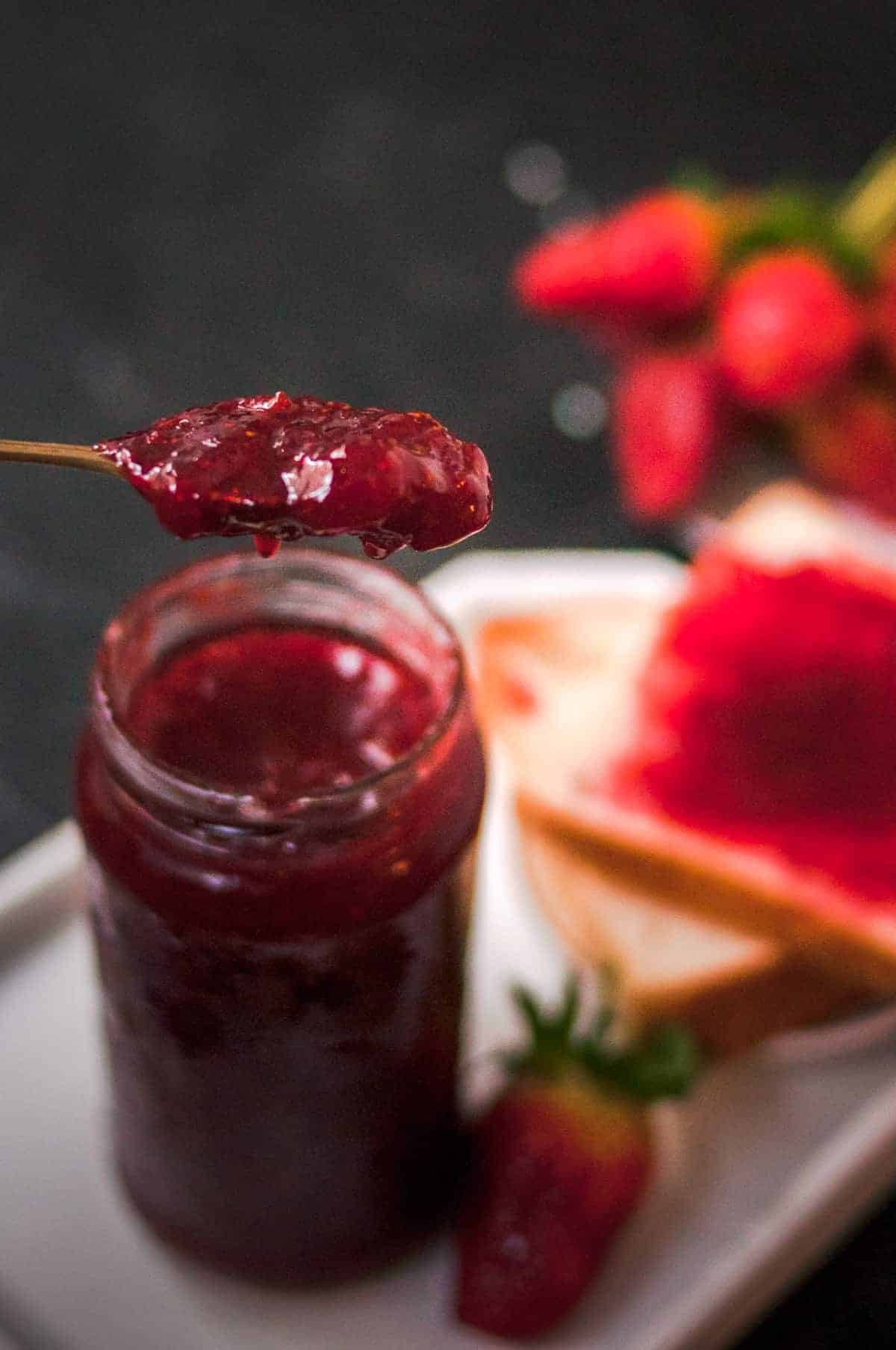 Strawberry jam jar on the black background with strawberries and jam toast behind the jar.