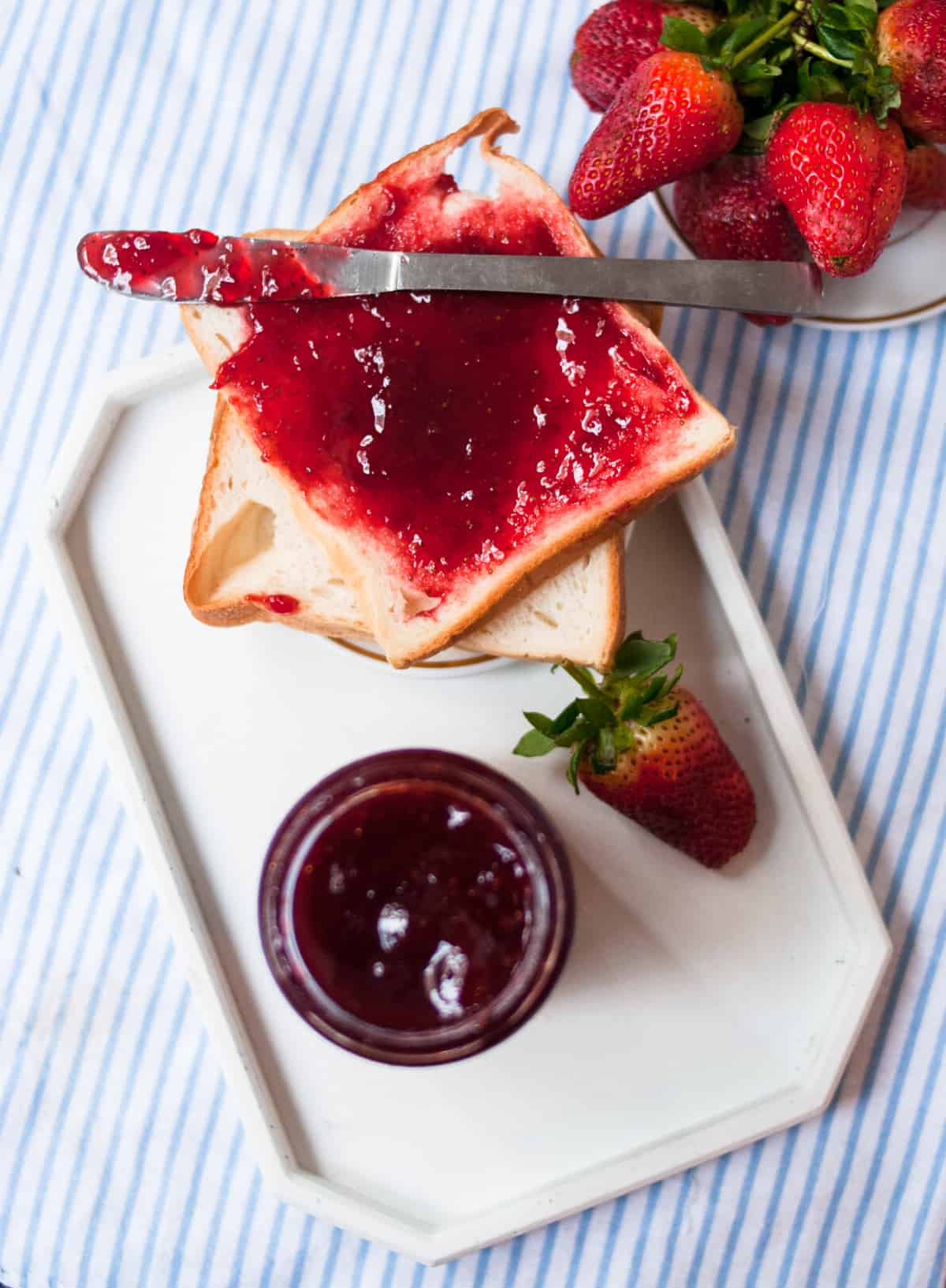 Strawberry jam on the bread slices with a butter knife over it on the blue striped kitchen towel.