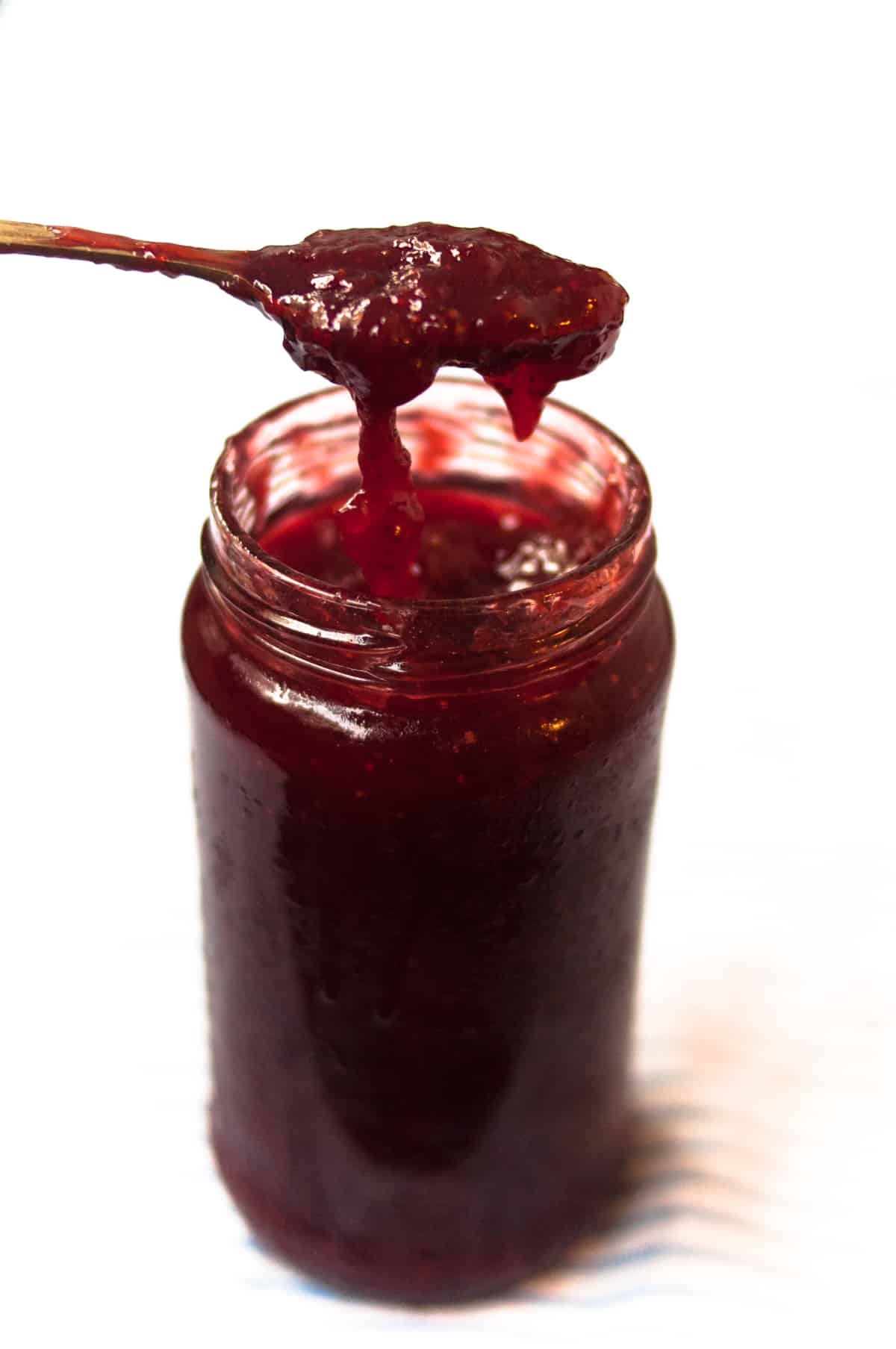 A jar of strawberry jam with a spoon of jam on it with jam overflowing.