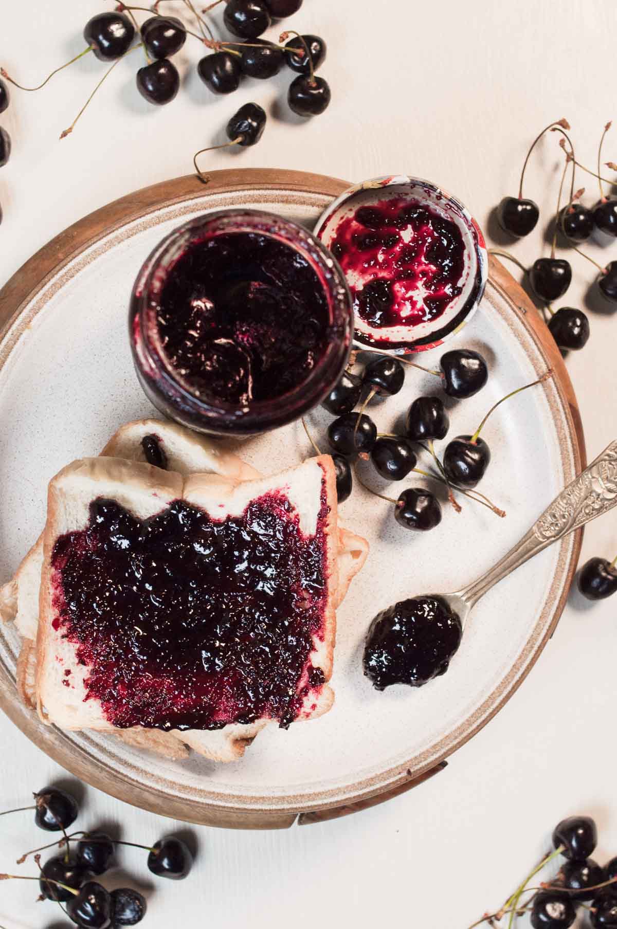 Cherry preserve in a jar with cherries all around and a lice of jam toast in the background.