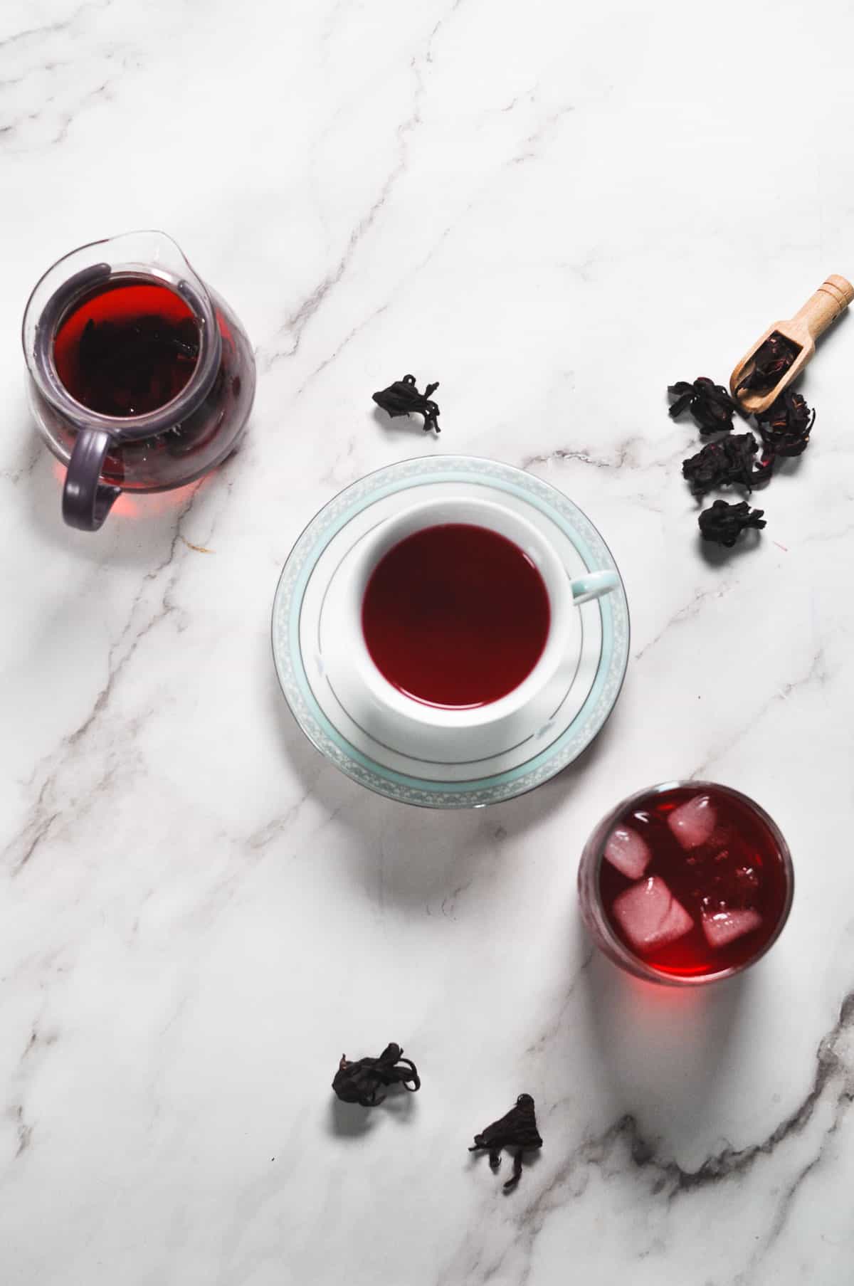 a kettle of steeped tea, a cup of hot tea and a glass of chilled iced with dried hibiscus flowers scattered on the white background.