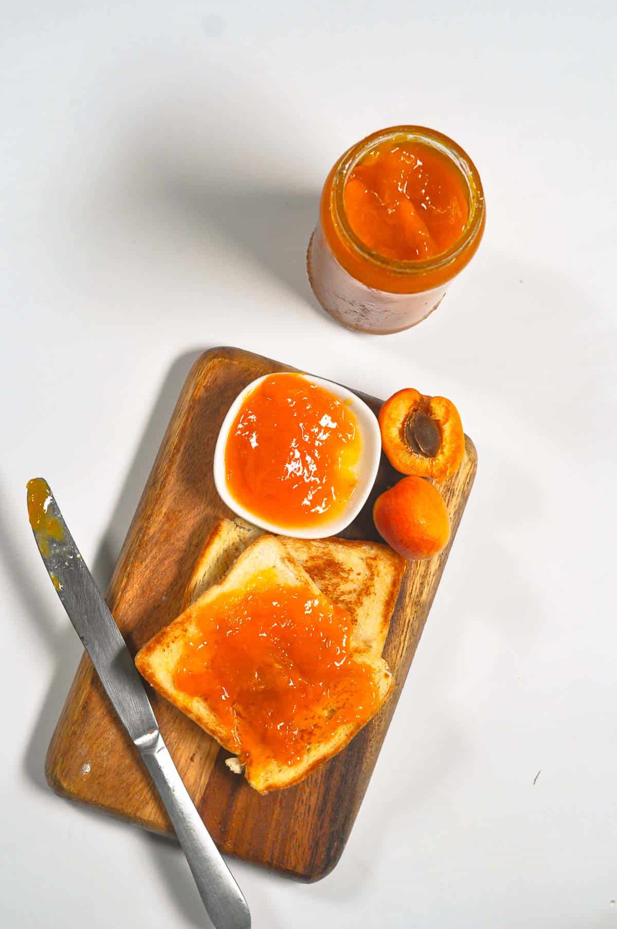 

Apricot jam in a glass jar and slathered on bread on a wooden cutting board.