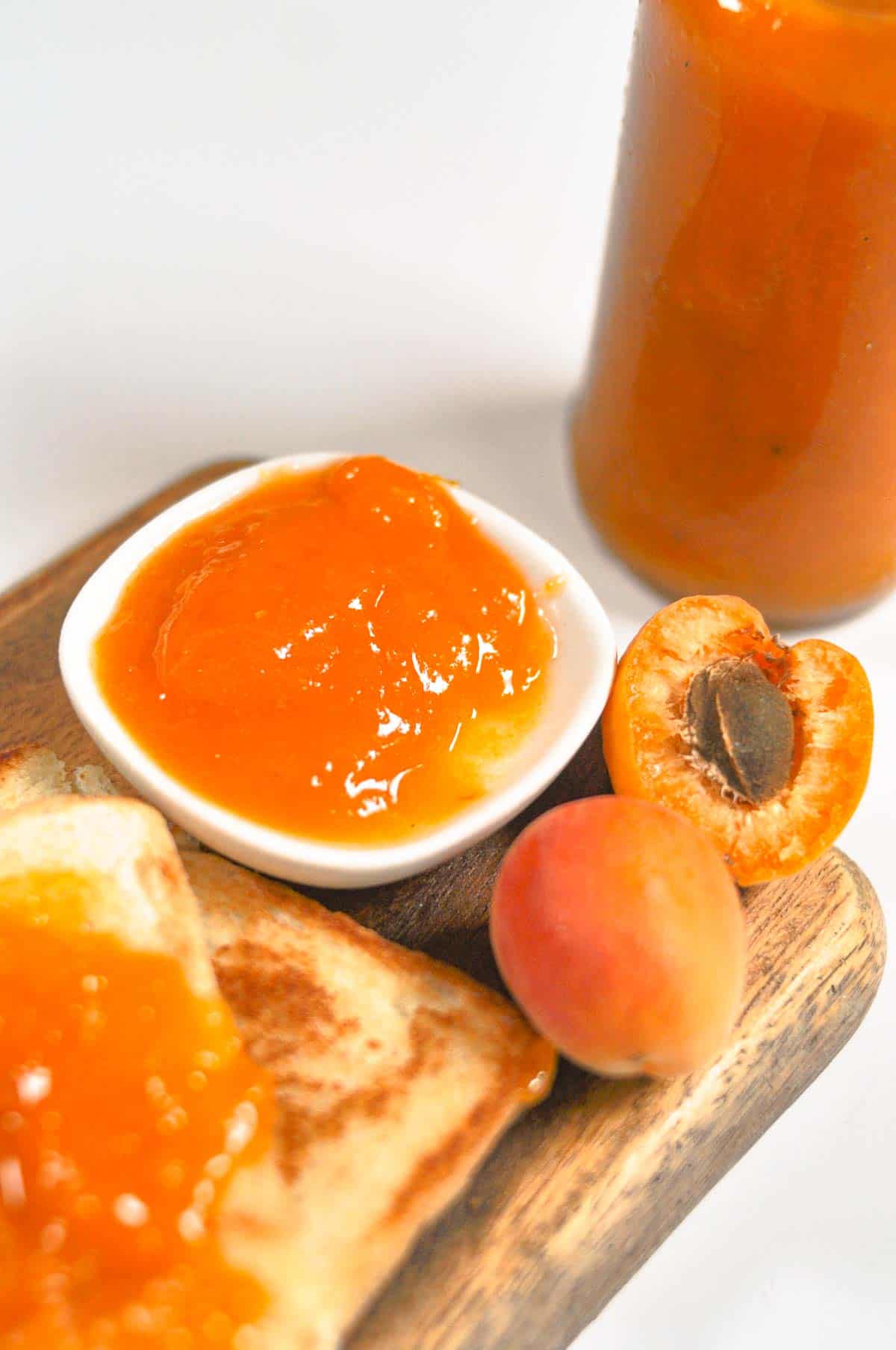 Apricot jam in a small plate is also spread on bread placed on a wooden cutting board.