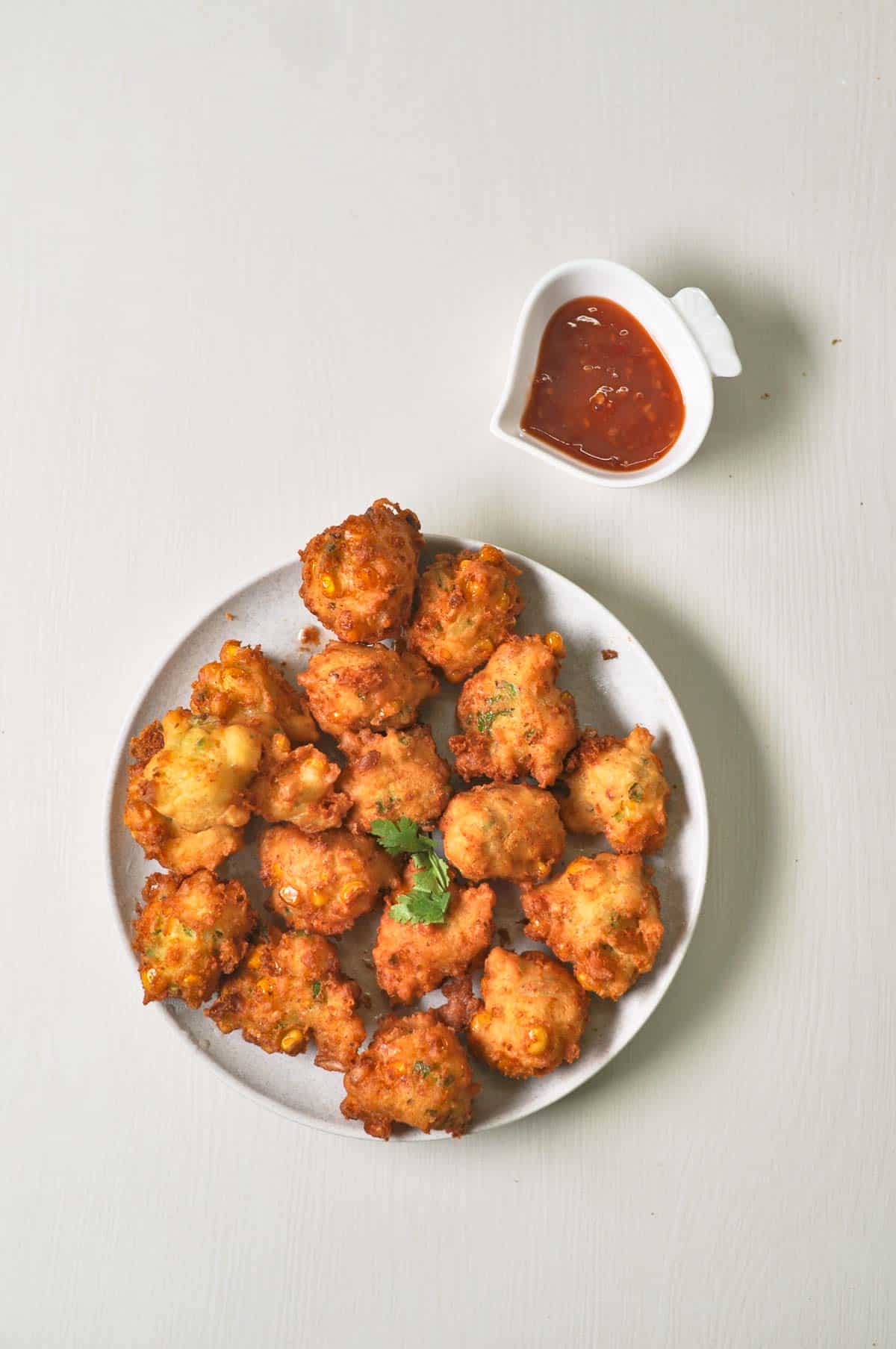 A white platter filled with golden-brown, crispy corn nuggets garnished with a sprig of cilantro, served with a small side bowl of red sweet chili sauce.