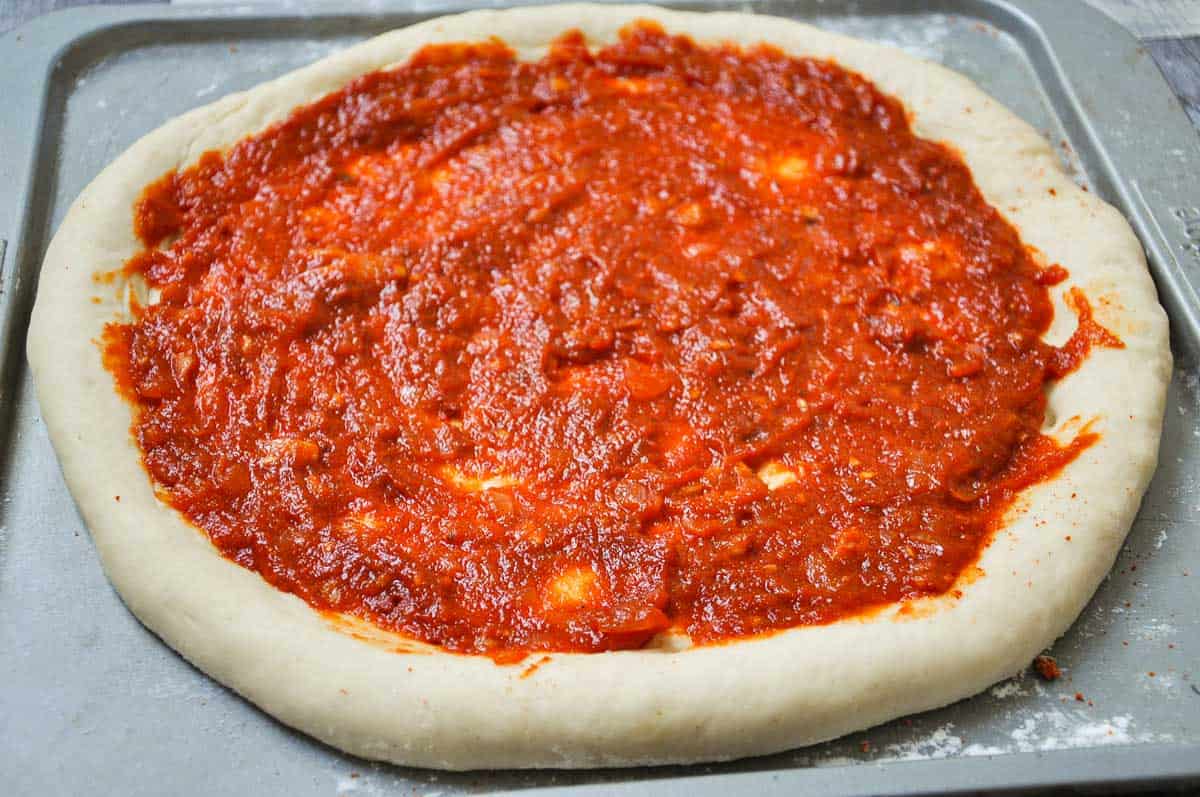 Pizza dough flattened on a tray, with pizza sauce spread over the top.