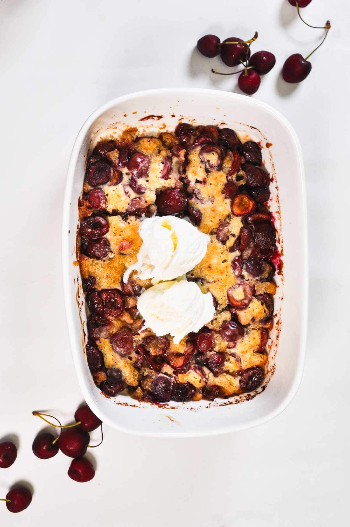 Overhead shot of baked cherry cobbler in a white rectangular dish, topped with two scoops of melting vanilla ice cream, surrounded by fresh cherries on a white marble surface.